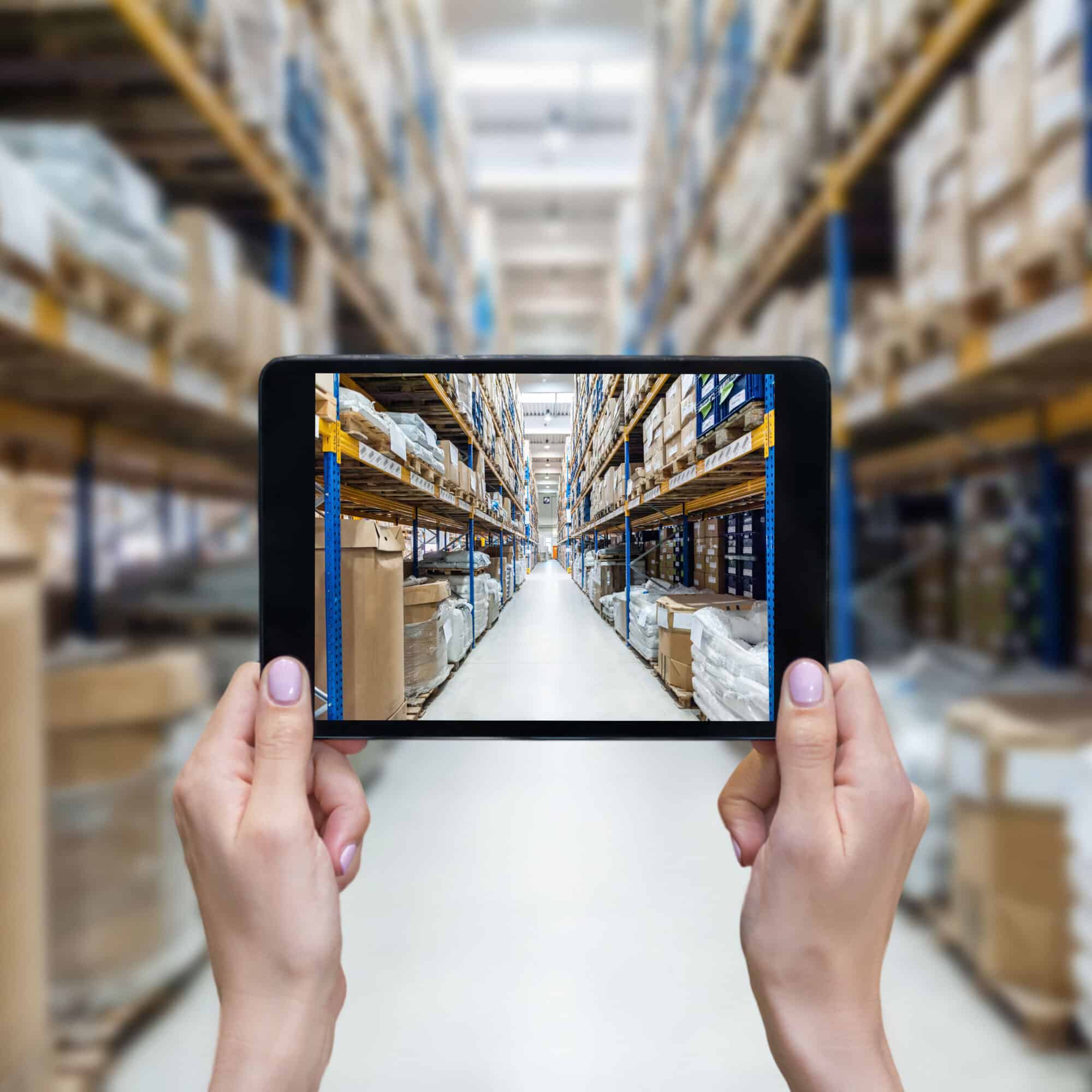 Horizontal color image of female hands holding a digital tablet in a corridor of futuristic distribution warehouse. Ordering on-line from a modern warehouse on a touchscreen tablet computer. Large distribution storage in background with racks full of packages, boxes, pallets, crates ready to be delivered. Logistics, freight, shipping, receiving.
